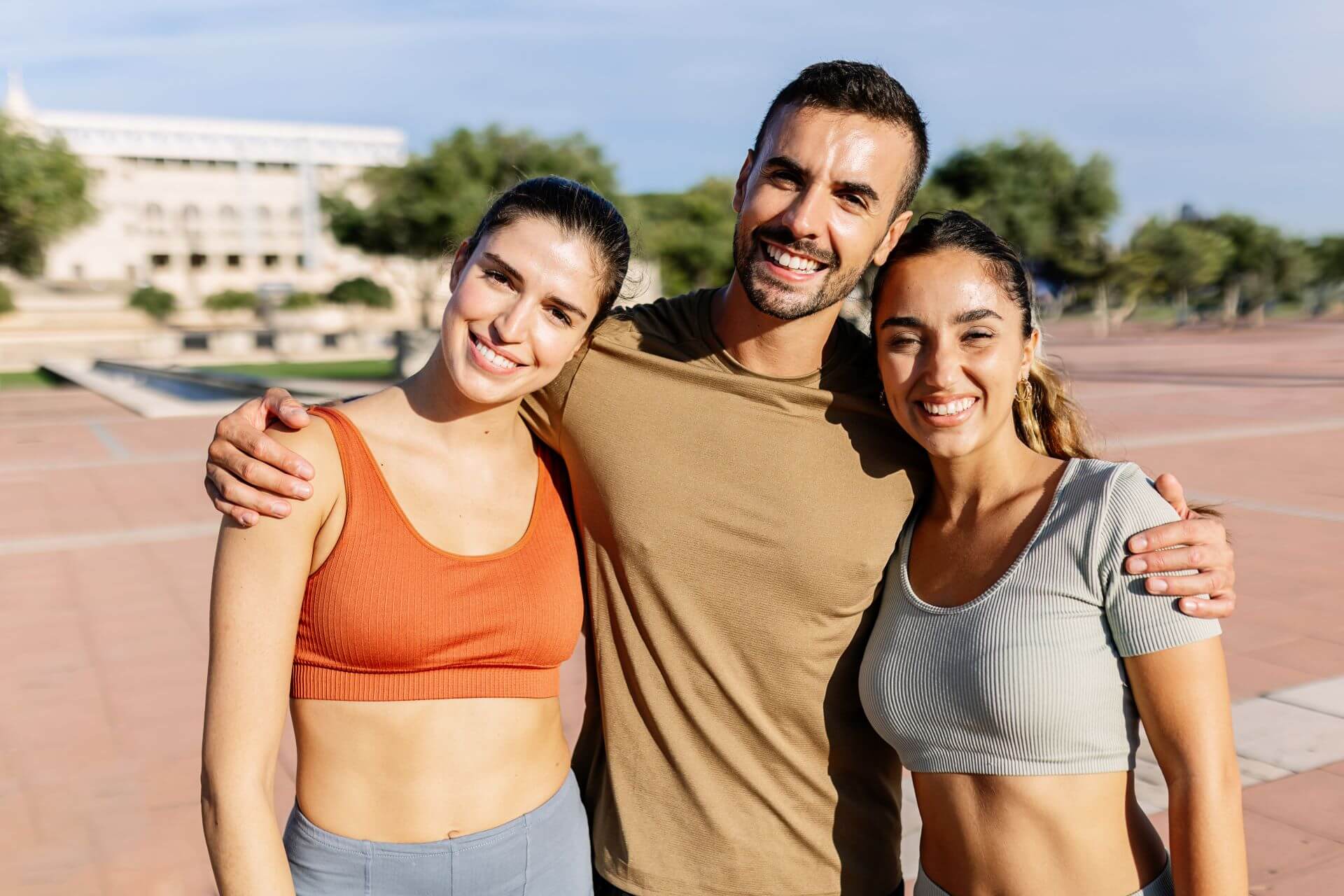 Three smiling people in workout gear stand together, one man and two women. They are outside on a sunny day.