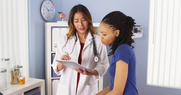 A woman doctor with a stethoscope reviews notes on a clipboard while speaking with a patient in a medical exam room.