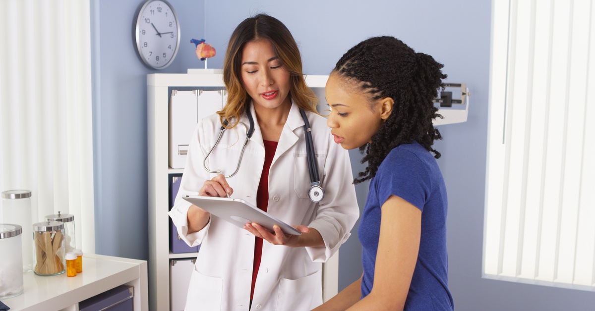 A woman doctor with a stethoscope reviews notes on a clipboard while speaking with a patient in a medical exam room.