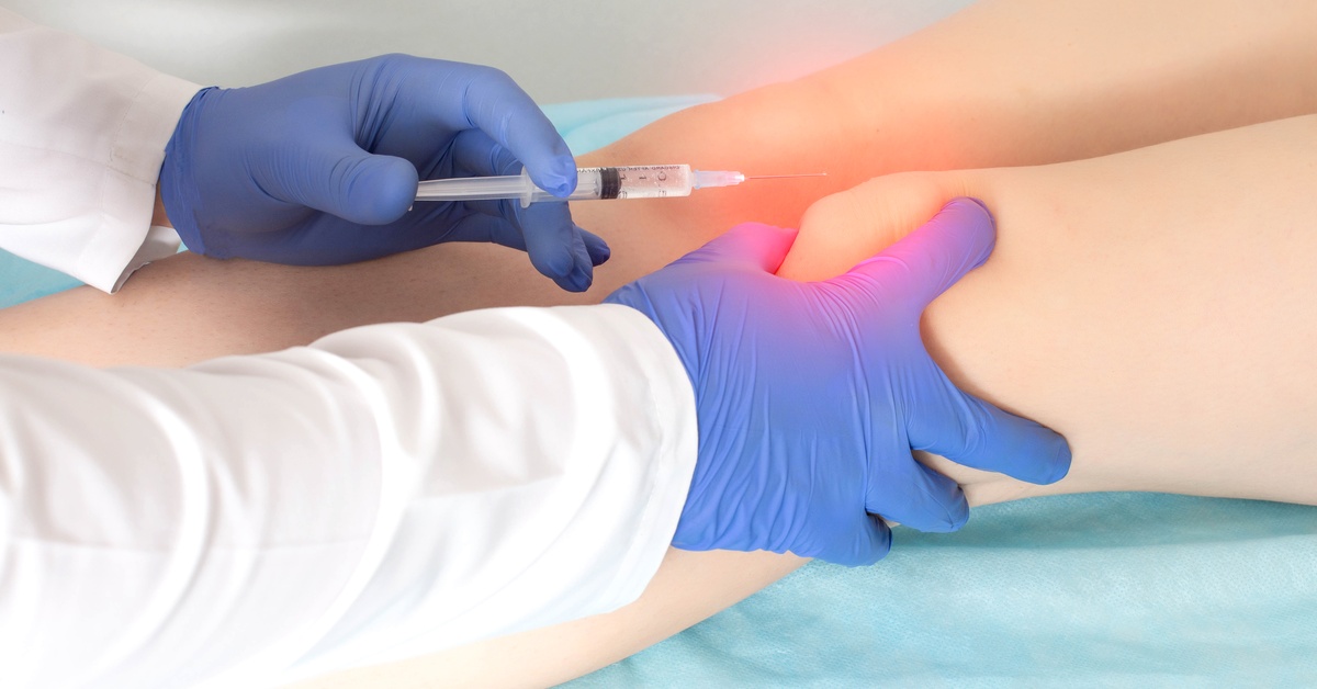 Gloved hands inject a syringe into a patient’s knee while applying pressure to the joint in a clinical setting.