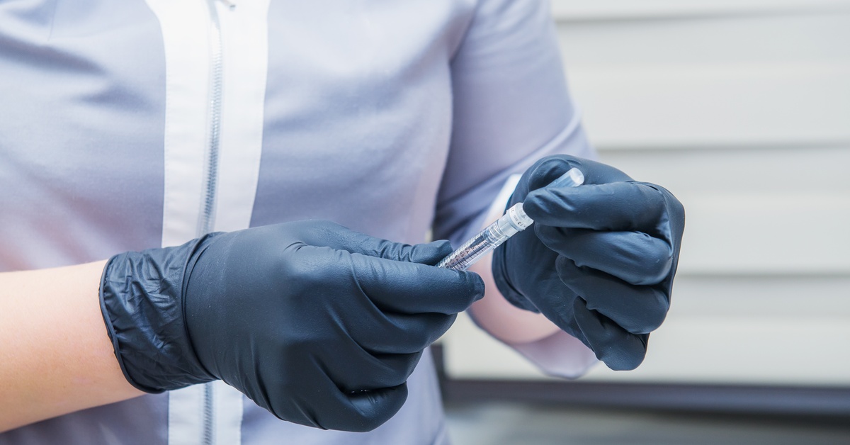 Gloved hands hold a syringe while preparing an injection, with a person wearing medical attire in the background.