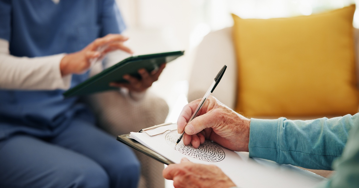 An older adult writes on a clipboard while a healthcare provider uses a tablet during a consultation session.