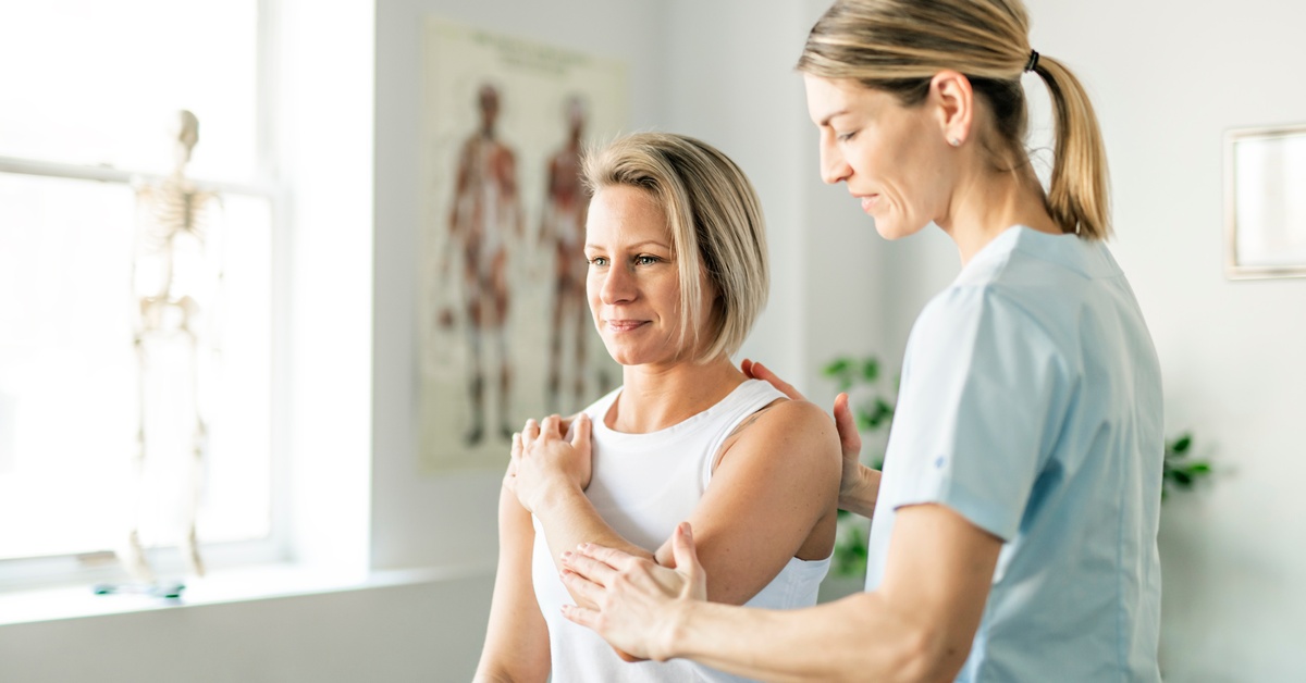 A clinician assesses a woman’s shoulder mobility during a physical exam in a bright, modern medical office.