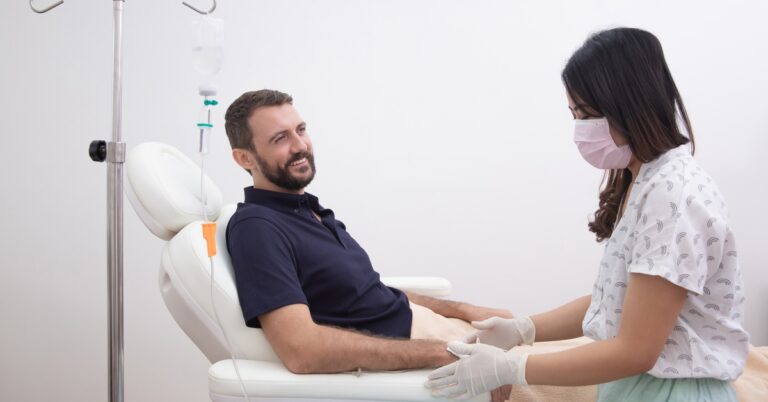 A patient receives IV therapy while a clinician monitors the infusion and adjusts the patient’s arm during treatment.