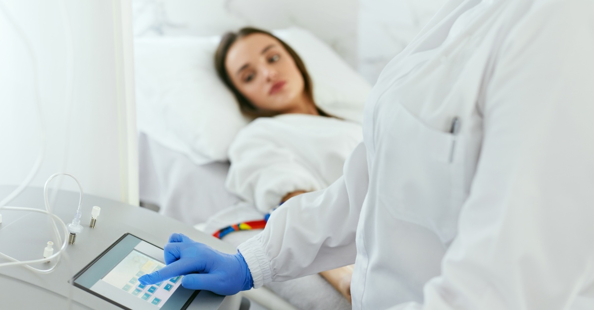 A clinician uses a touchscreen device while a patient lies in a medical bed receiving treatment in a clinical setting.