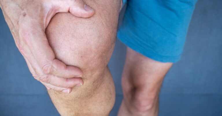 A person grips their knee with both hands, suggesting pain or discomfort in the joint while standing.