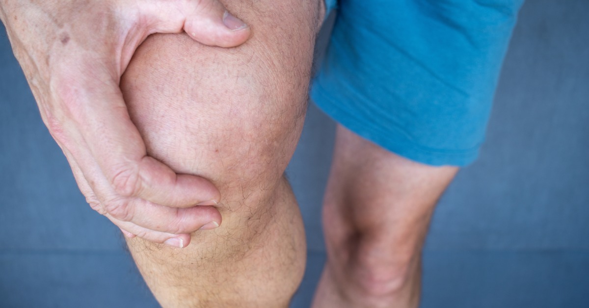 A person grips their knee with both hands, suggesting pain or discomfort in the joint while standing.
