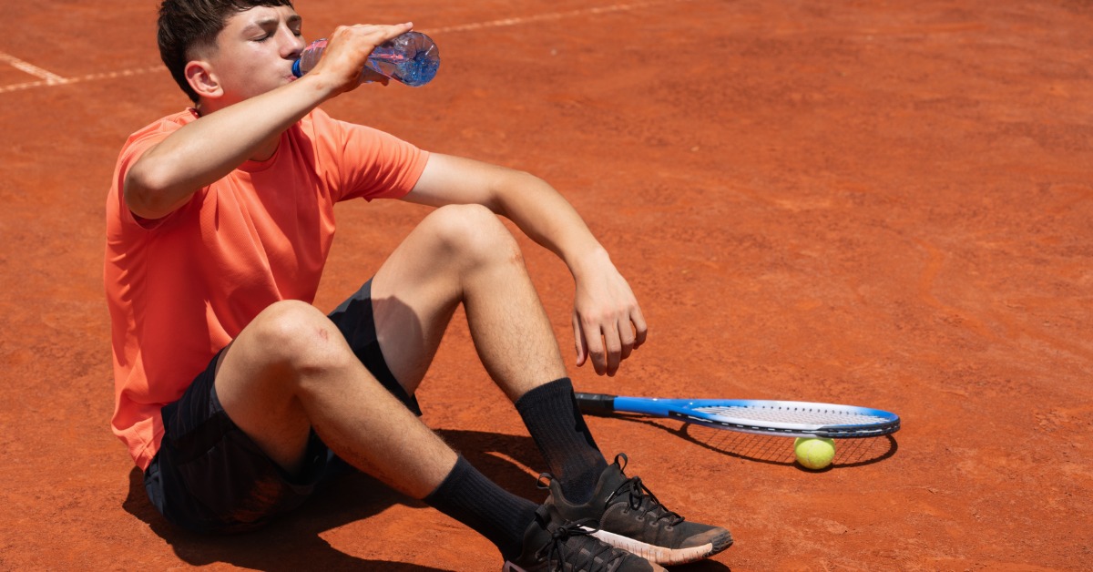 A tennis player sits on a clay court drinking water during a break, with a racket and ball resting nearby.