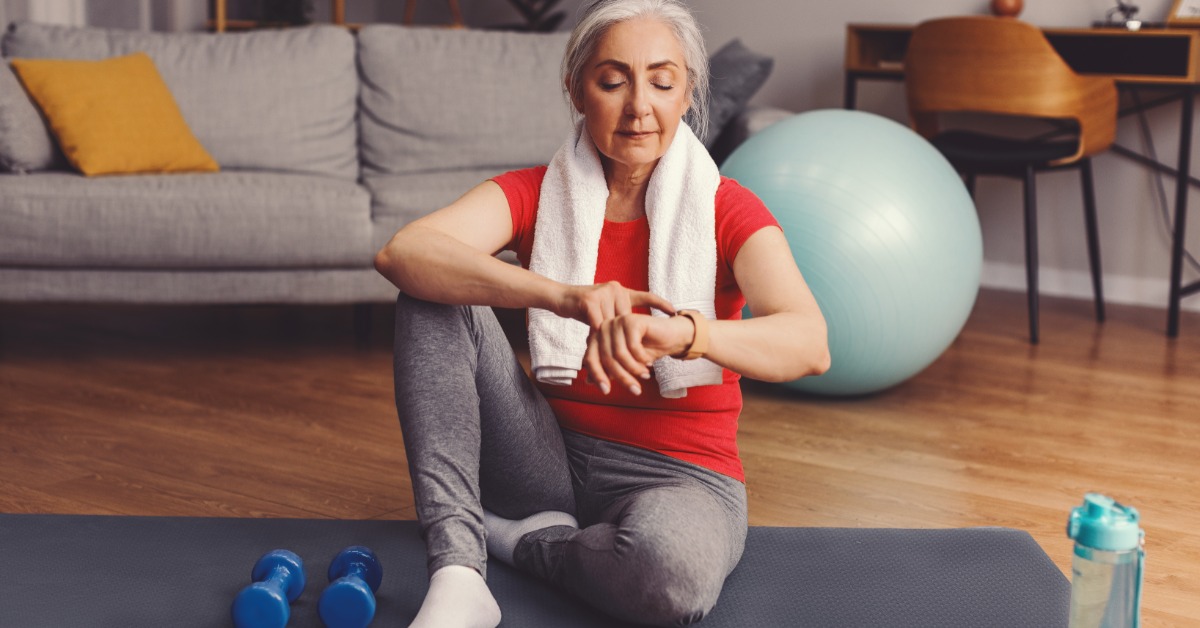 An older woman sits on a yoga mat checking her smartwatch after a workout, with dumbbells and a water bottle nearby.