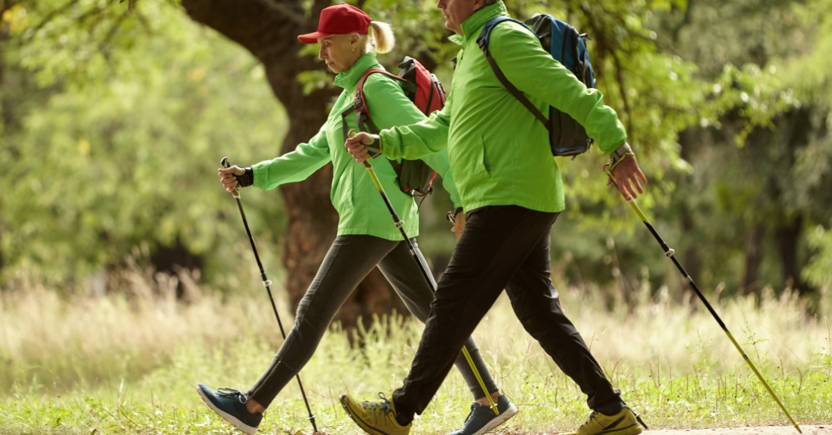 Two older adults use walking poles while hiking together on a wooded trail as part of an active outdoor lifestyle.