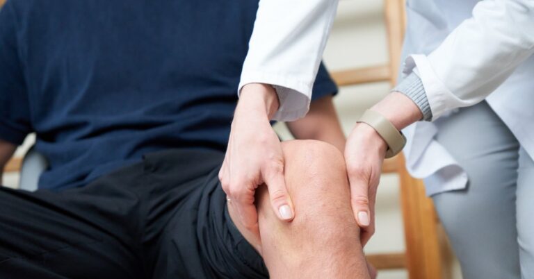A healthcare professional in a white coat holds a seated man's knee with both hands during an examination.