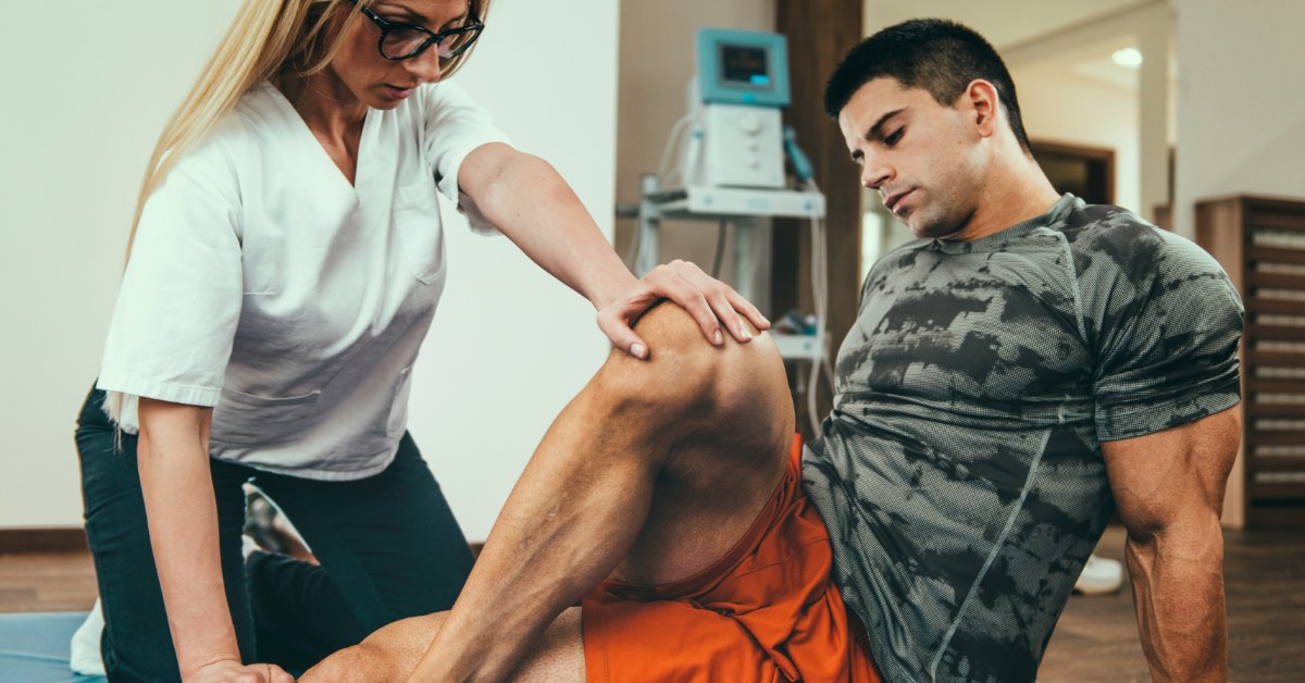 A female healthcare professional holds a muscular man's bent knee and ankle as he sits on an exercise mat on the floor.