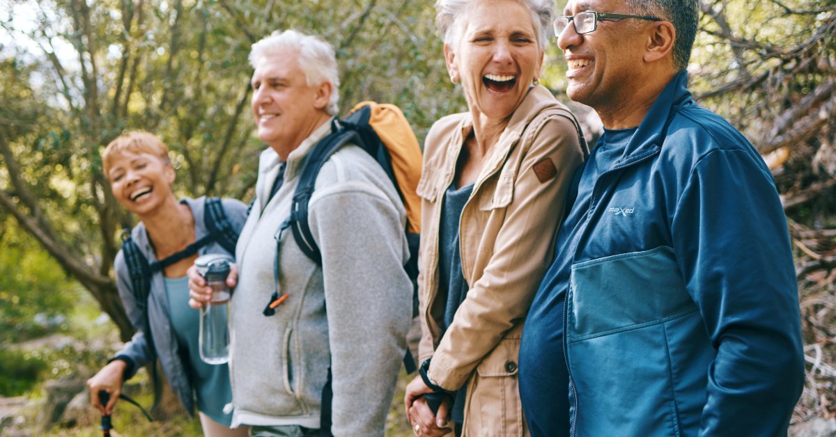 Four older adults enjoy a hike together on a wooded trail while carrying walking poles and outdoor gear.