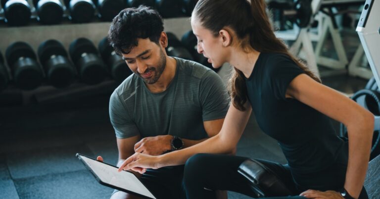 A man and a woman review health information together on a clipboard while sitting in a fitness facility.