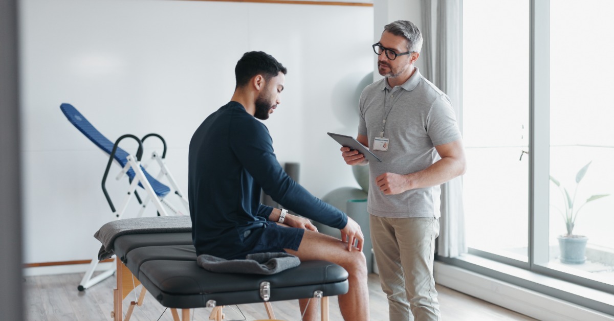A patient speaks with a healthcare professional holding a tablet while seated on a treatment table for a consultation.